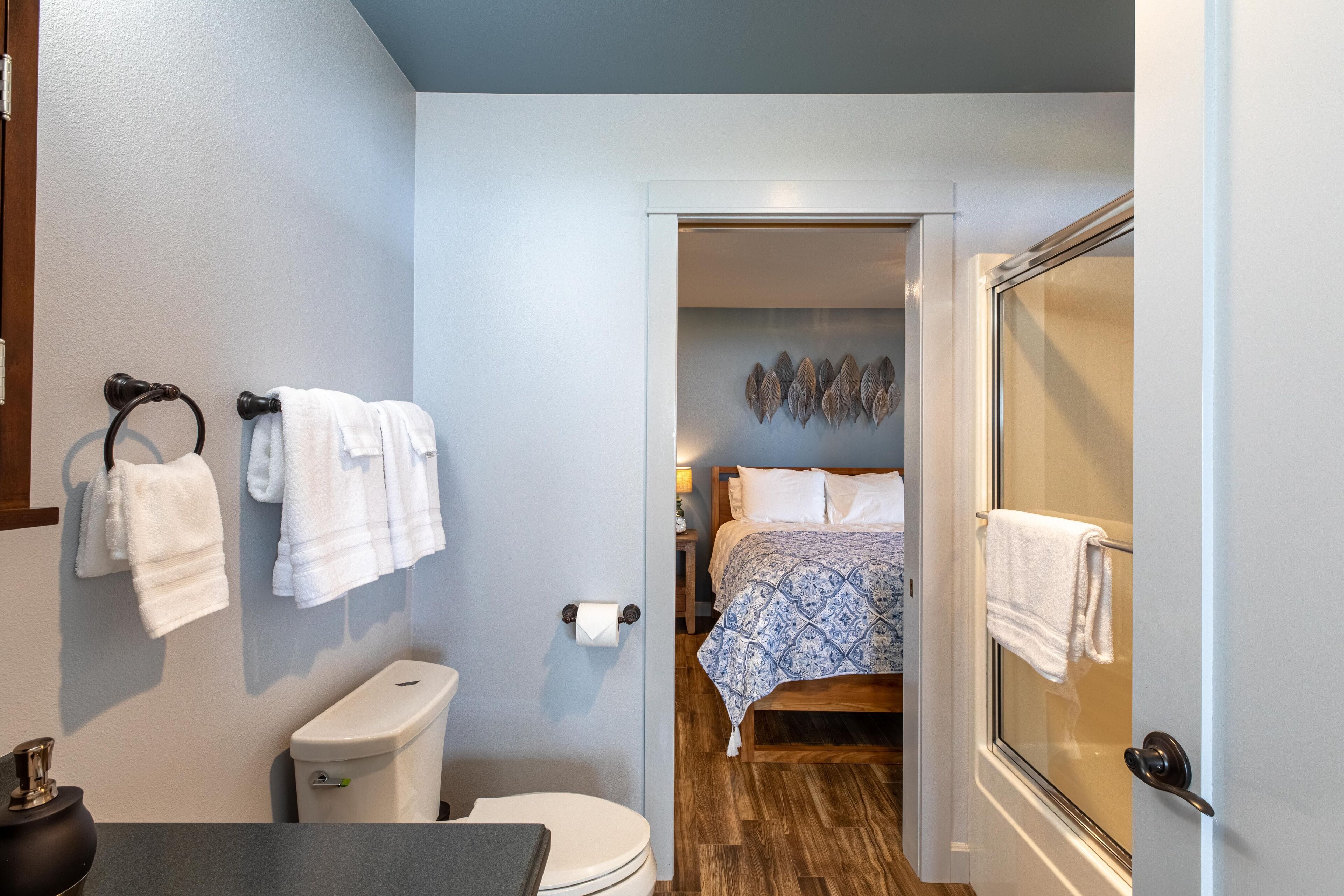 Un baño con inodoro, lavabo y barra para toallas. Se ve un dormitorio a través de una puerta entreabierta, que incluye una cama y una mesita de noche.