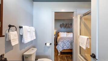 Un baño con inodoro, lavabo y barra para toallas. Se ve un dormitorio a través de una puerta entreabierta, que incluye una cama y una mesita de noche.