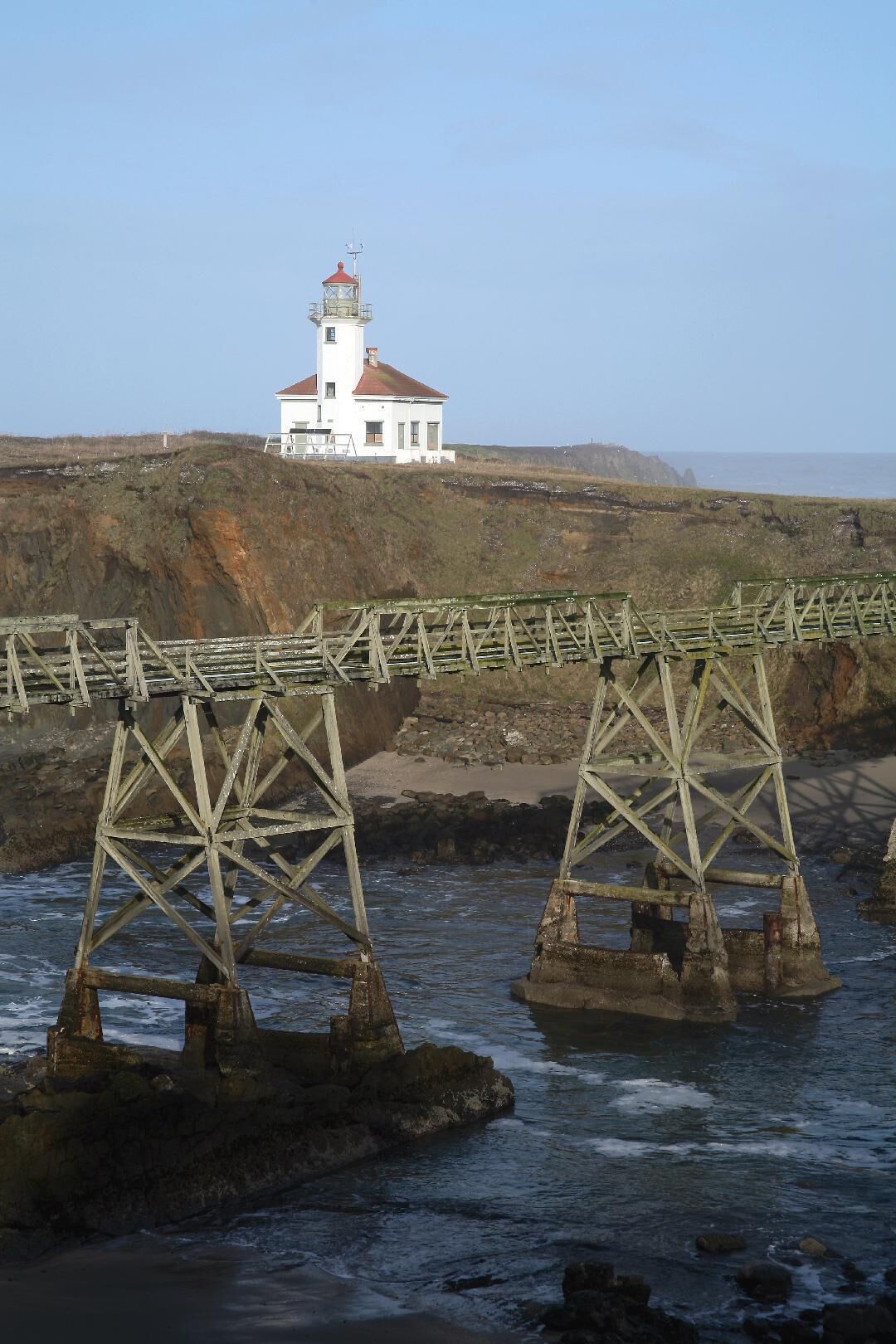 A white lighthouse with a red roof on a cliff overlooking the sea, with a wooden bridge in the foreground.
