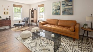 Ein Wohnzimmer mit einer Ledersofa, einem Glastisch, einer Holzkommode und zwei Jalousiefenstern.