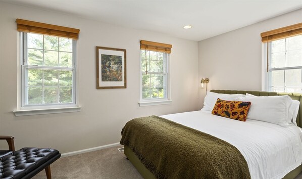A bedroom with a large bed, two windows with blinds, a framed picture on the wall, and a bedside lamp.