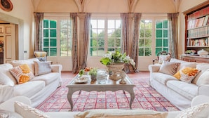 Amplia habitación con dos sofás blancos, una mesa de centro y una estantería. Grandes ventanales con contraventanas verdes dejan entrar luz natural y ofrecen vistas al exterior.
