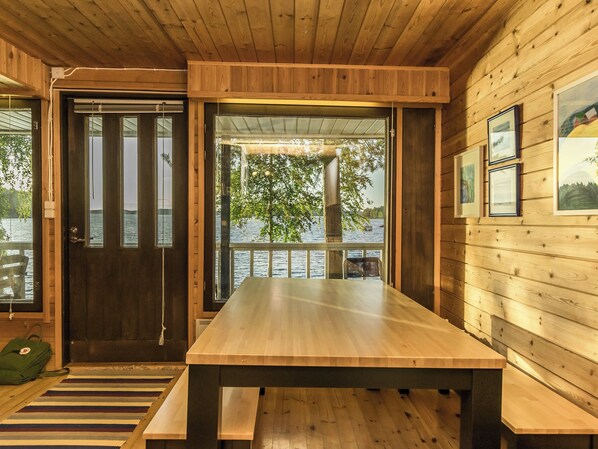 A wooden cabin interior with a large table, a door leading outside, and framed pictures on the wall.