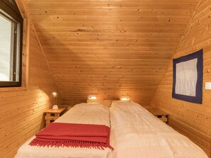 A small, cozy room with wooden walls and a sloped ceiling, featuring two beds with white bedding and a red blanket, a small table, and a wall-mounted light fixture. - Kiviranta by Interhome (Savonlinna)