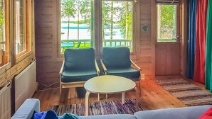 Une pièce chaleureuse avec un plancher en bois, un canapé, deux fauteuils, une petite table et de grandes fenêtres donnant sur des arbres et une étendue d’eau.