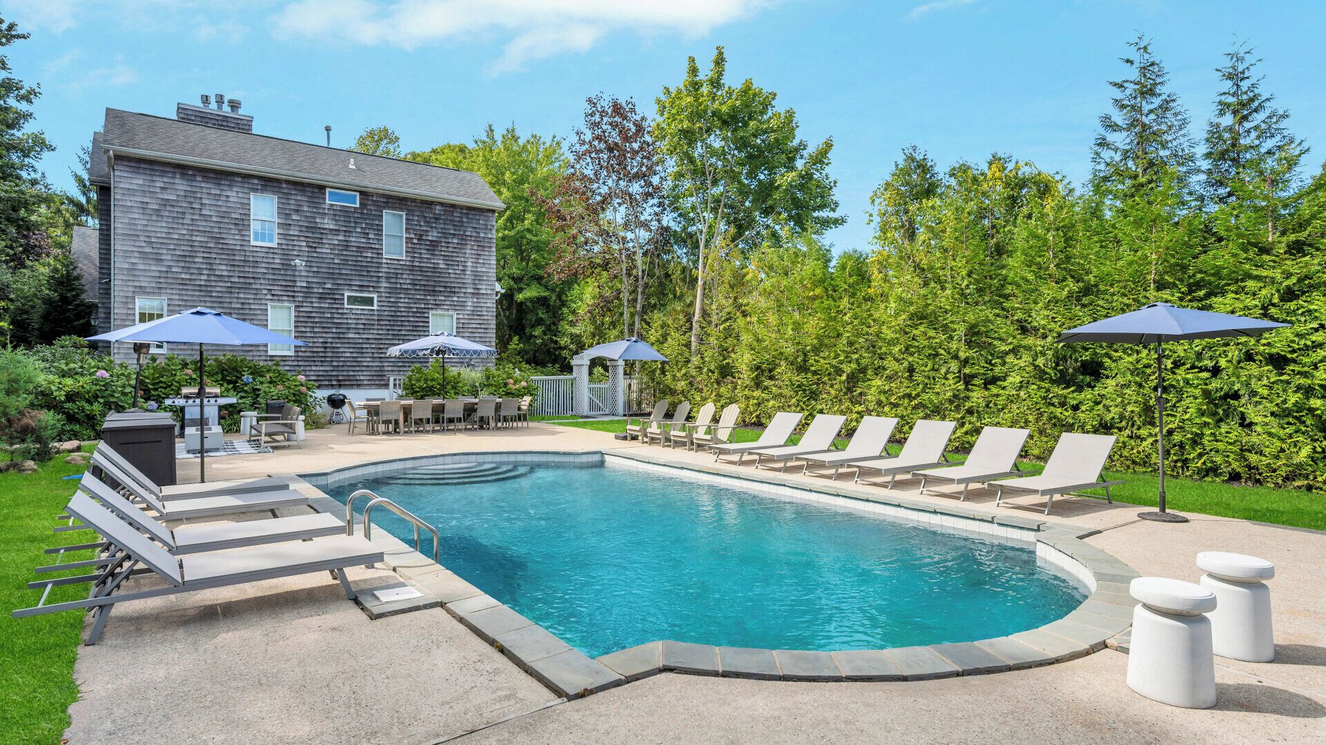 A swimming pool with lounge chairs and umbrellas, a barbecue area, and a house in the background.