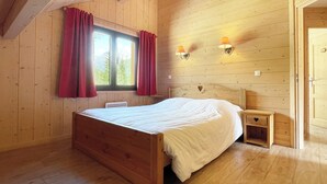 A wooden room with a bed, two windows with curtains, a bedside table, and wall-mounted lights. - BOURG D'OISANS/ALPE d'HUEZ ★ CHALET Bois & Nature with panoramic view (LE BOURG D'OISANS)