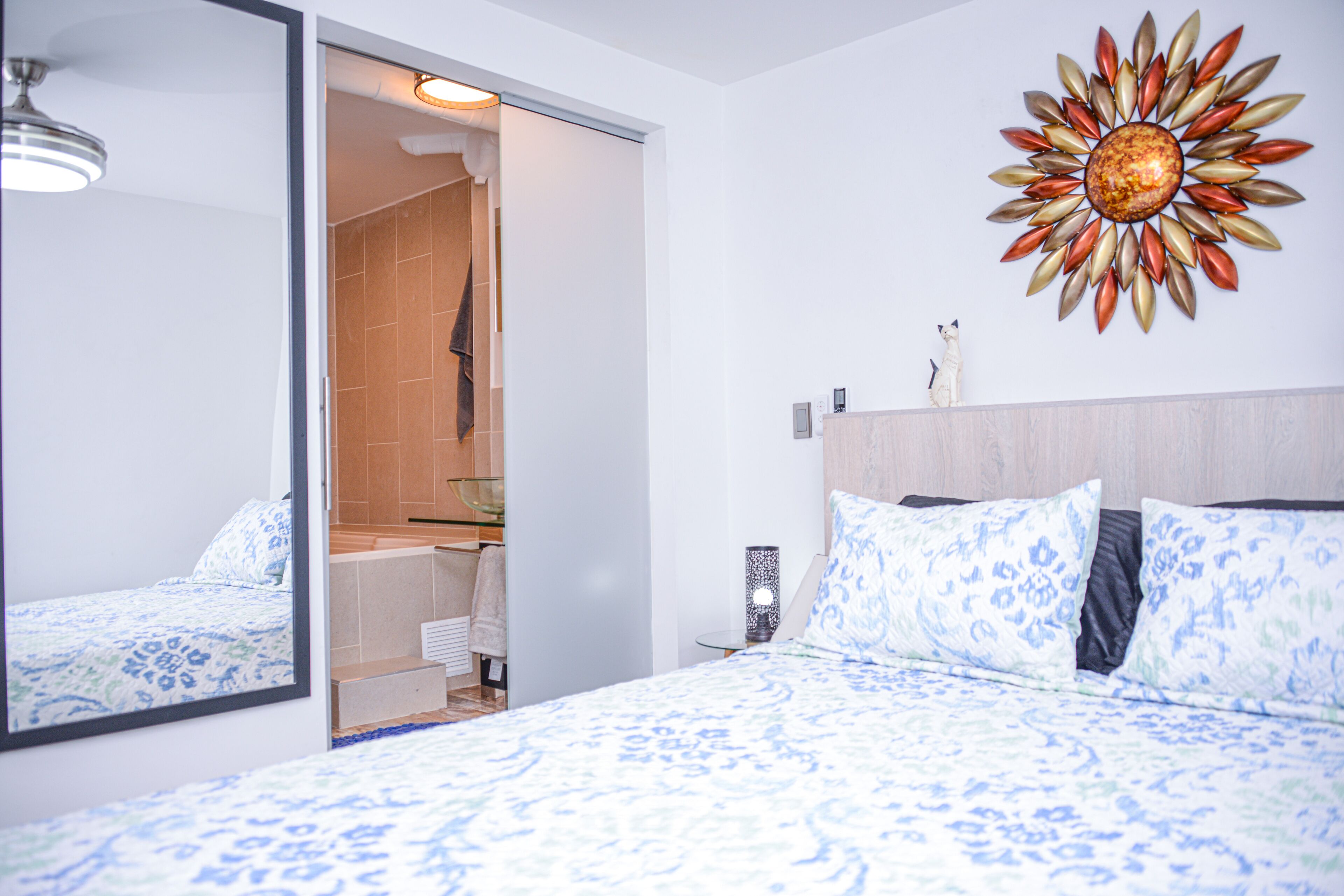Een moderne slaapkamer met een groot bed, een badkamer zichtbaar door een geopende deur, en een decoratief zonvlecht muurkunstwerk.