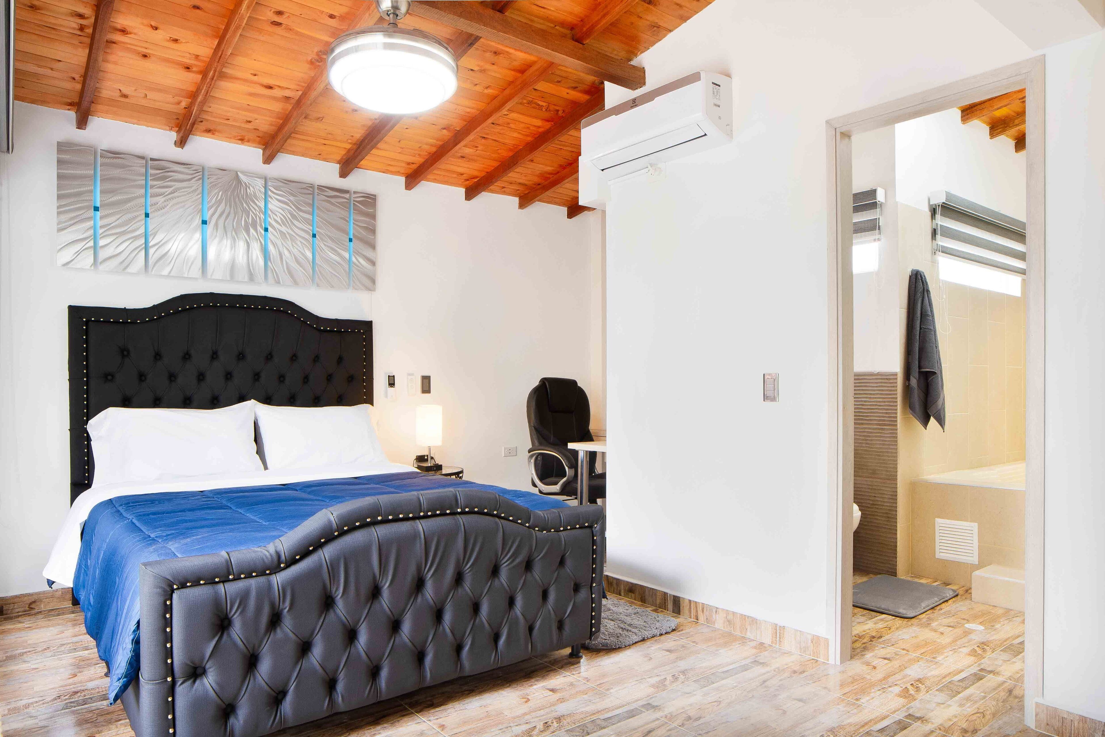 A bedroom with a large bed, a wooden ceiling, an air conditioning unit, and a bathroom visible through an open door.