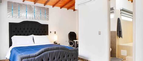 A bedroom with a large bed, a wooden ceiling, an air conditioning unit, and a bathroom visible through an open door.