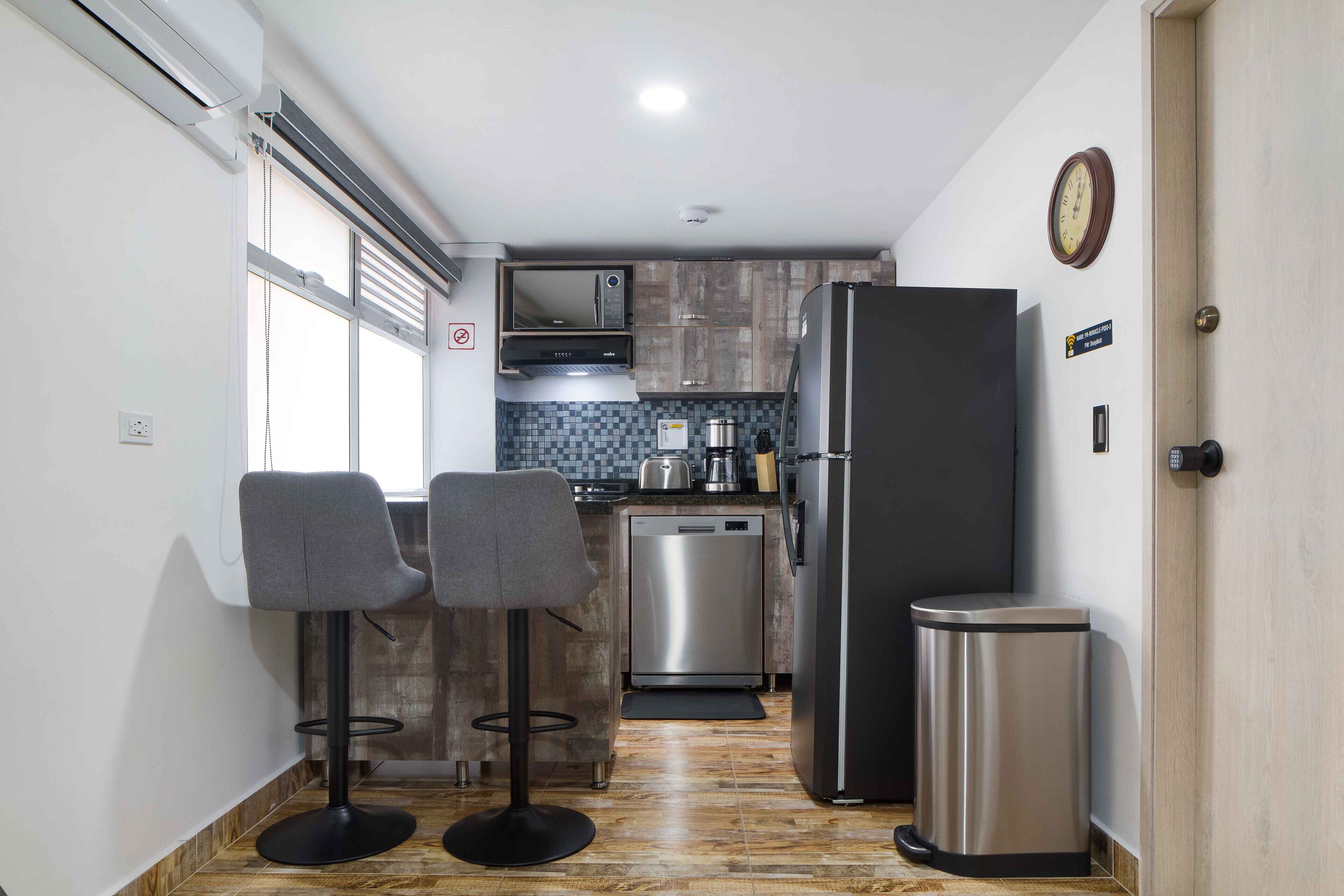 A compact kitchen with a refrigerator, microwave, and dishwasher. There are two bar stools and a small trash bin.