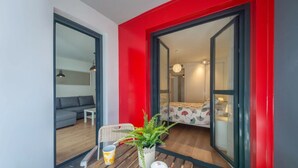 Een modern appartement met een rode accentmuur, een balkon met een rieten stoel, een klein tafeltje met een plant en een mok, en een slaapkamer met een bed en een nachtkastje.