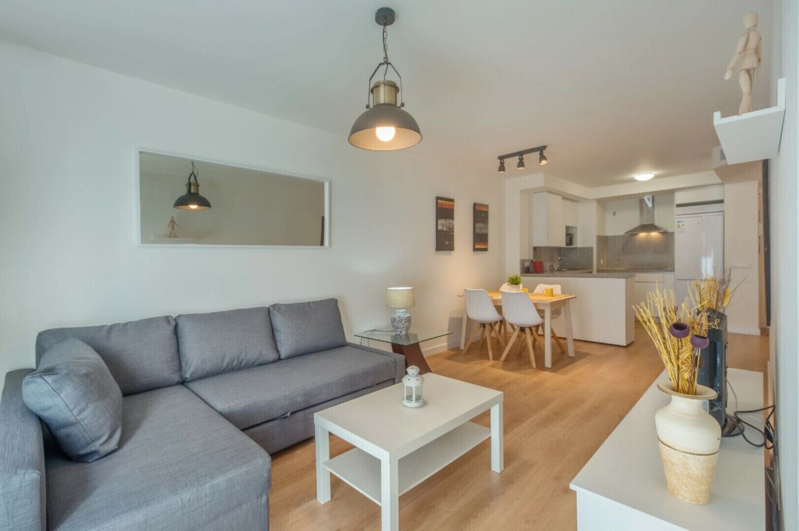 Uma sala de estar moderna com um sofá cinzento, uma mesa de centro branca e uma área de jantar com uma pequena mesa e cadeiras. Uma cozinha é visível ao fundo.