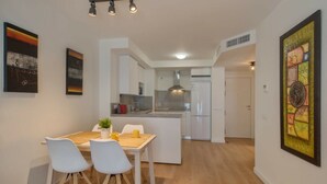Een moderne keuken met witte kasten, een ingebouwde oven en een koelkast. Er is een eethoek met een witte tafel en stoelen, en kunstwerken aan de wanden.