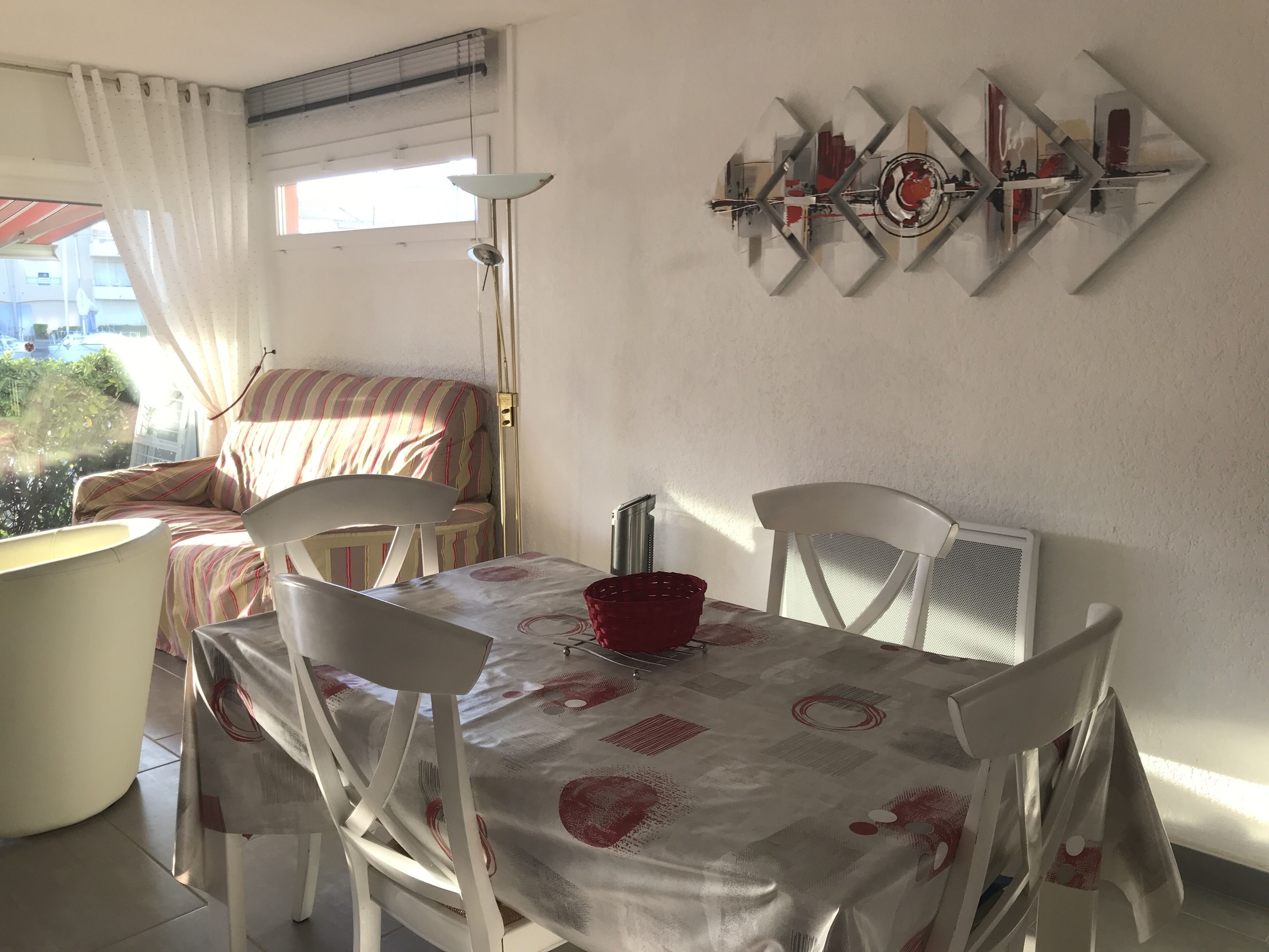 A dining area with a table set for two, white chairs, a red bowl, and a wall art piece.