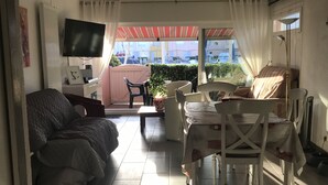 A compact living space with a sofa, a dining table with chairs, a TV mounted on the wall, and a view of the outside through a large window. - PRETTY GROUND FLOOR CONTEMPORARY GARDEN BROWN (Cap d'Agde)