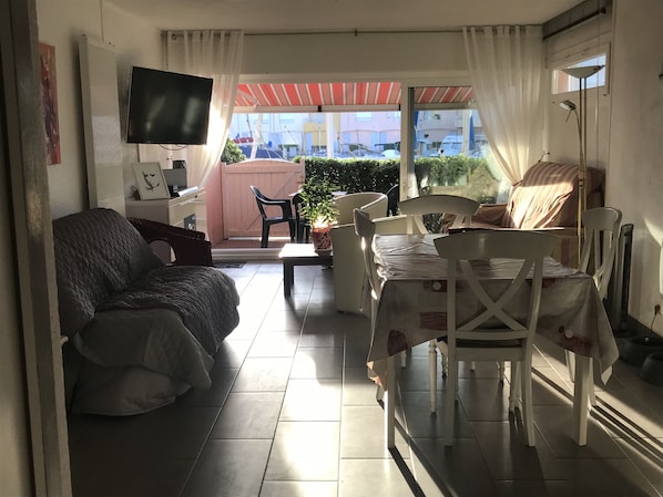 A compact living space with a sofa, a dining table with chairs, a TV mounted on the wall, and a view of the outside through a large window. - PRETTY GROUND FLOOR CONTEMPORARY GARDEN BROWN (Cap d'Agde)