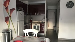 A compact kitchen with a refrigerator, microwave, and stove. There's a dining table with a red basket and a white chair. A clock is on the wall. - PRETTY GROUND FLOOR CONTEMPORARY GARDEN BROWN (Cap d'Agde)