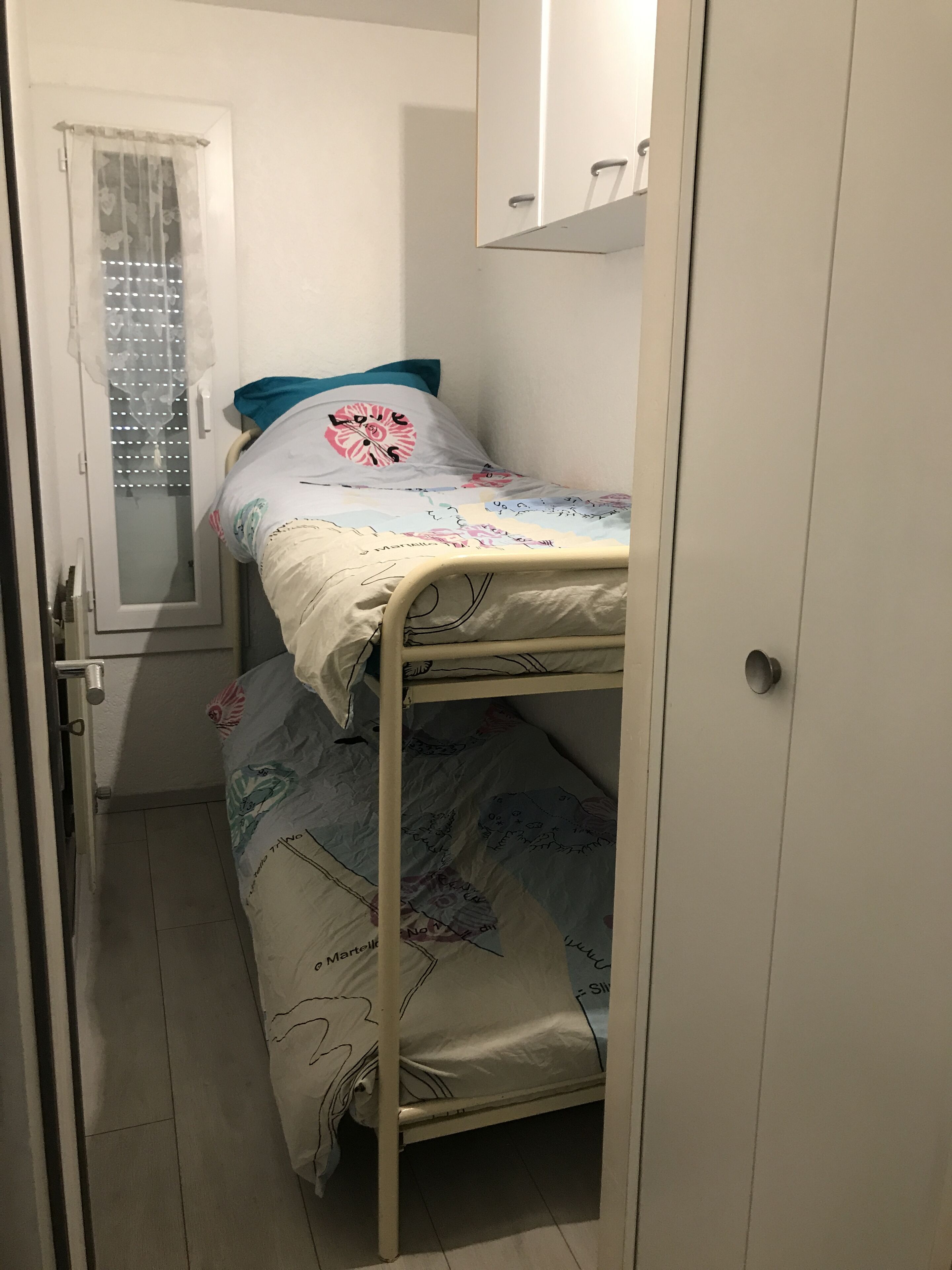 Una habitació petita i estreta amb un llit superposat, una porta amb un finestral motllurat i un armari blanc.