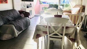 A room with a sofa, a dining table with chairs, a TV, and a view of the outside. - PRETTY GROUND FLOOR CONTEMPORARY GARDEN BROWN (Cap d'Agde)