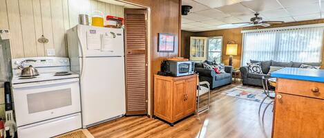 Cocina compacta con estufa, refrigerador y microondas. En la sala de estar, hay un mueble de madera, un televisor, un sofá y una mesa azul.