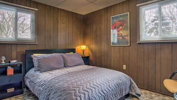 Un dormitorio con pared de paneles de madera, una cama con colcha estampada, una mesita de noche con lámpara y un cuadro en la pared.