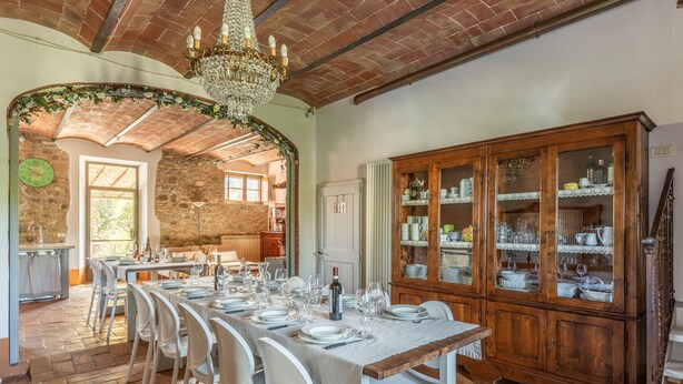 Uma sala de jantar com uma mesa comprida posta para refeição, rodeada por cadeiras brancas. Um armário de madeira com portas de vidro é visível à direita.