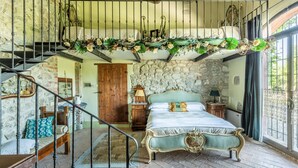 Quarto com parede de pedra, cama com cabeceira verde, um banco de madeira, escada com corrimão de ferro e vista para o exterior através de uma janela.