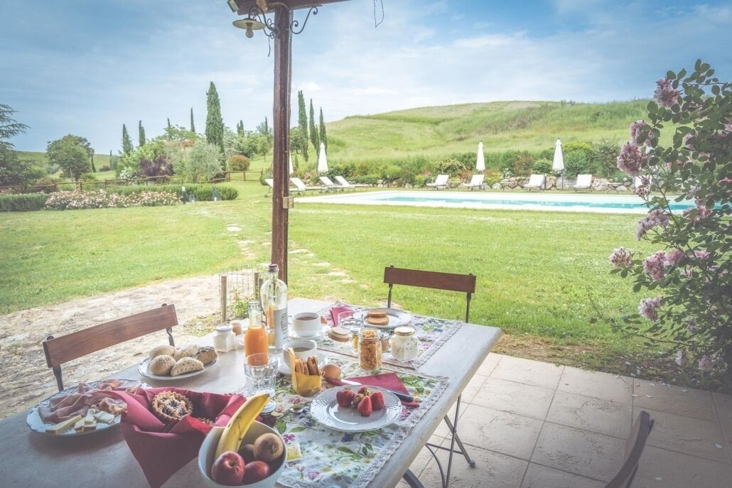 Un área de comedor al aire libre con una mesa preparada para el desayuno, con vista a una piscina y un paisaje cubierto de césped.
