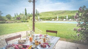 Uma área de refeições ao ar livre com uma mesa posta para café da manhã, com vista para uma piscina e paisagem gramada.