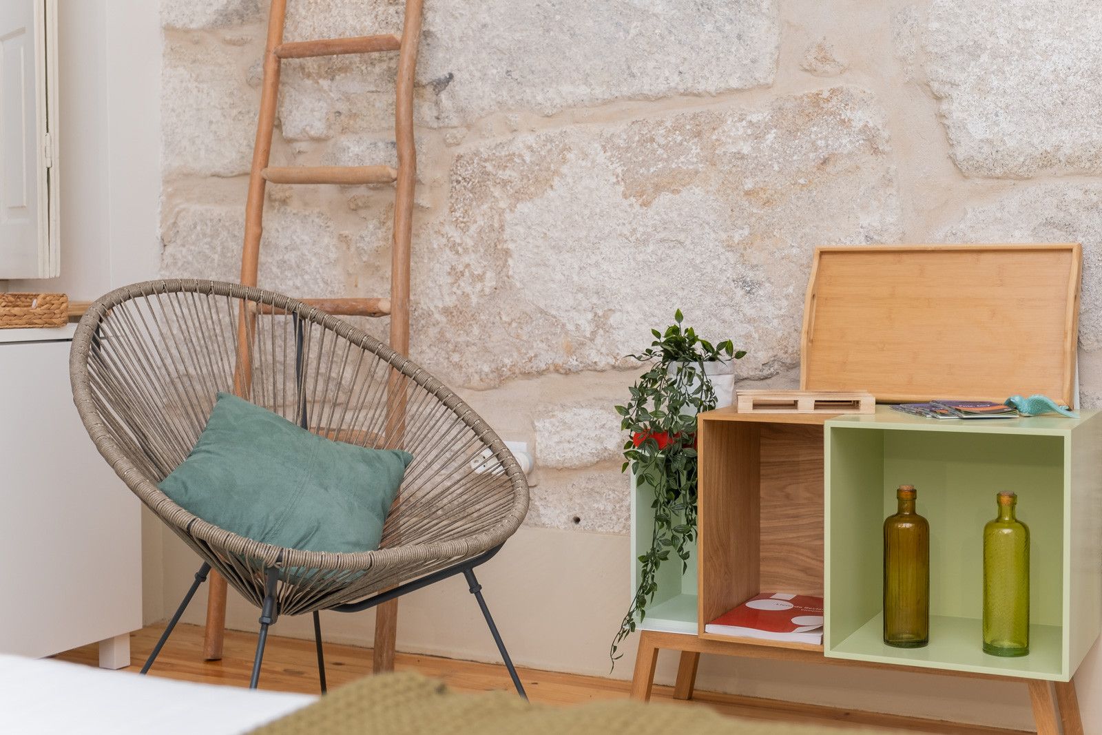 Une chaise en osier avec un coussin, une échelle en bois et une étagère verte sur laquelle se trouvent deux bouteilles et une plante.