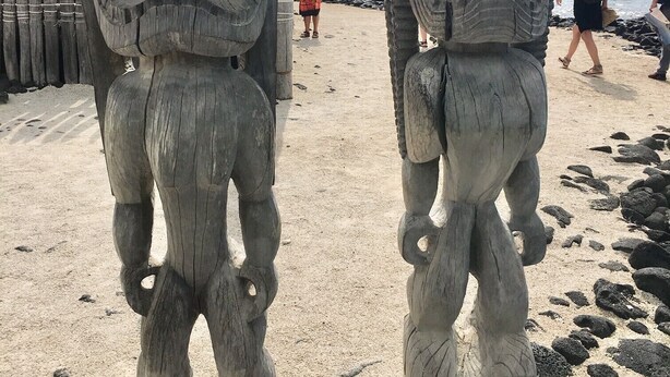 Dos grandes estatuas de tiki de piedra en una playa arenosa, con una costa rocosa al fondo.