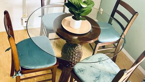 Un comedor con una mesa de vidrio rodeada de sillas de madera con asientos acolchados, una planta en maceta sobre la mesa y un televisor montado en la pared.