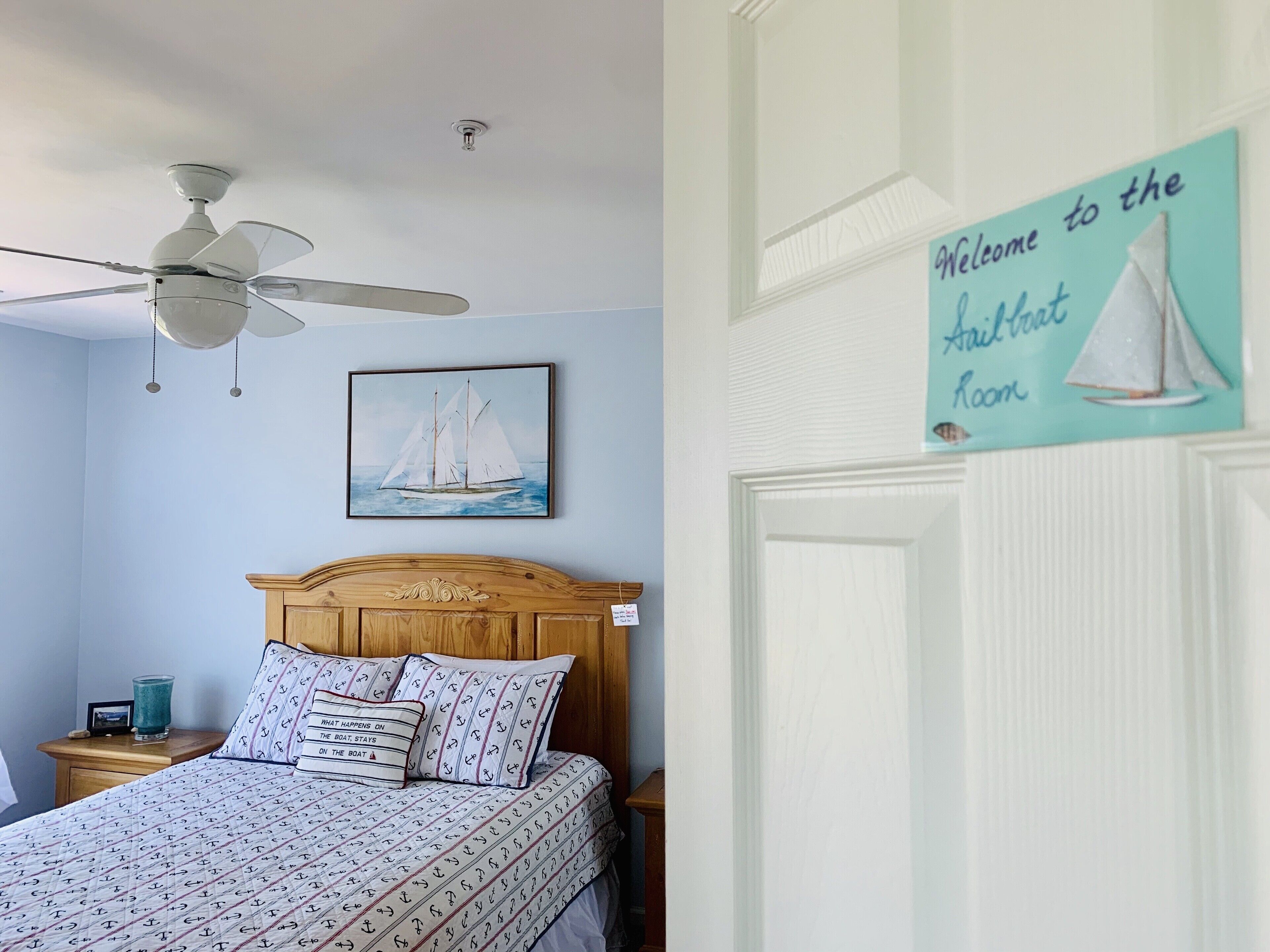 A bedroom with a bed, a ceiling fan, a sailboat painting, and a welcome sign.
