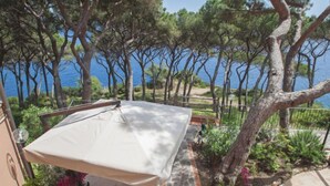A patio with a table and chairs under a canopy, surrounded by trees and a view of the sea.