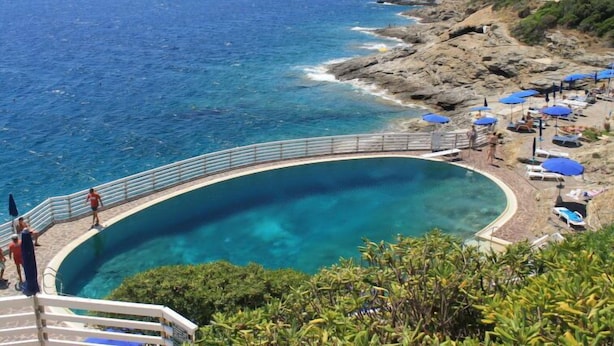 A curving pool with a clear view of the ocean, surrounded by a rocky coastline and lush greenery.