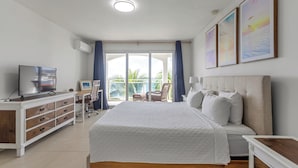 A hotel room with a large bed, a TV, a desk with a chair, and a balcony with a view of palm trees. - Maho Beach House: Sunset Suite, 1-Bedroom (Maho)