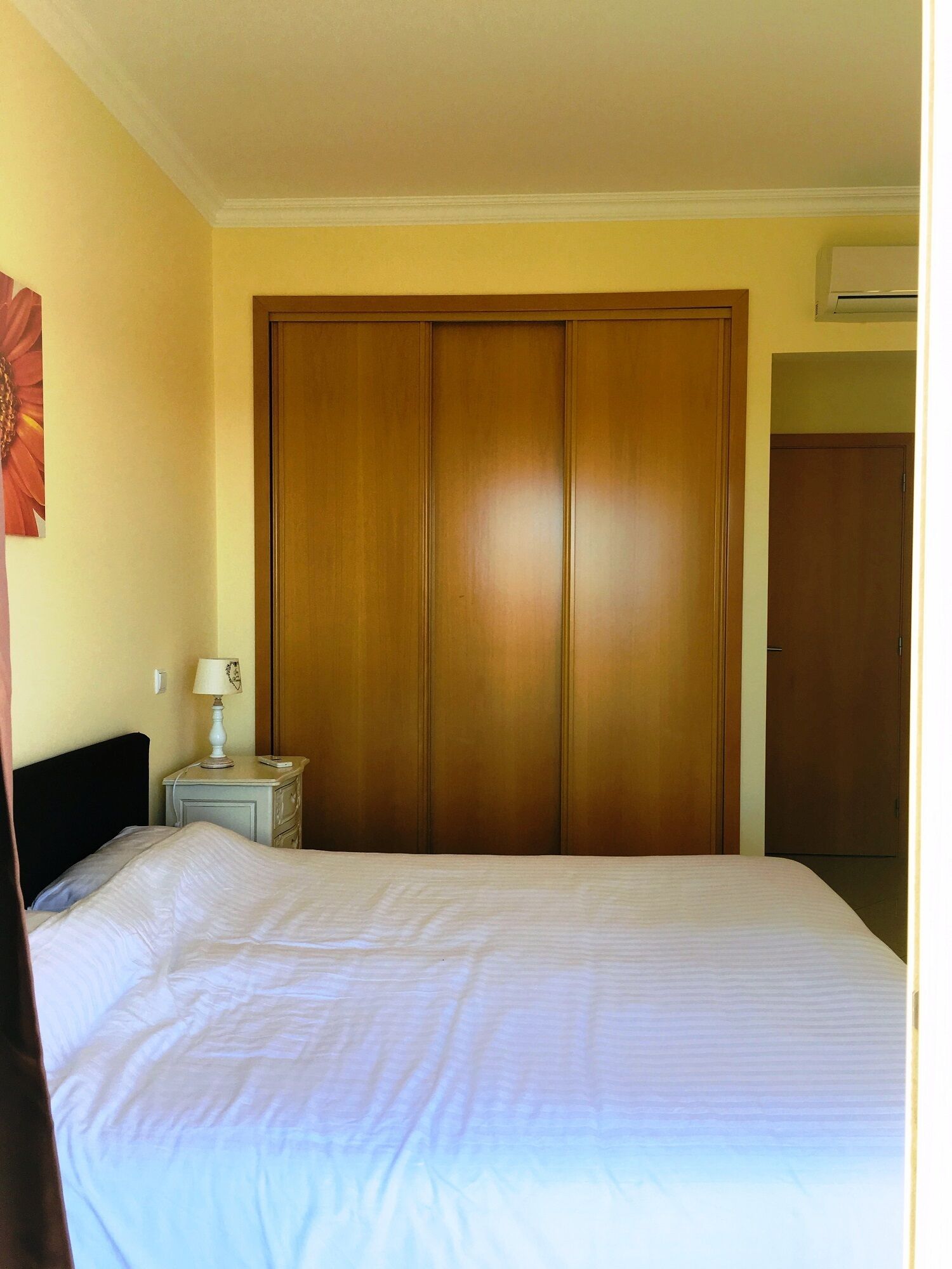 A bedroom with a large bed, a nightstand with a lamp, and a wooden wardrobe.