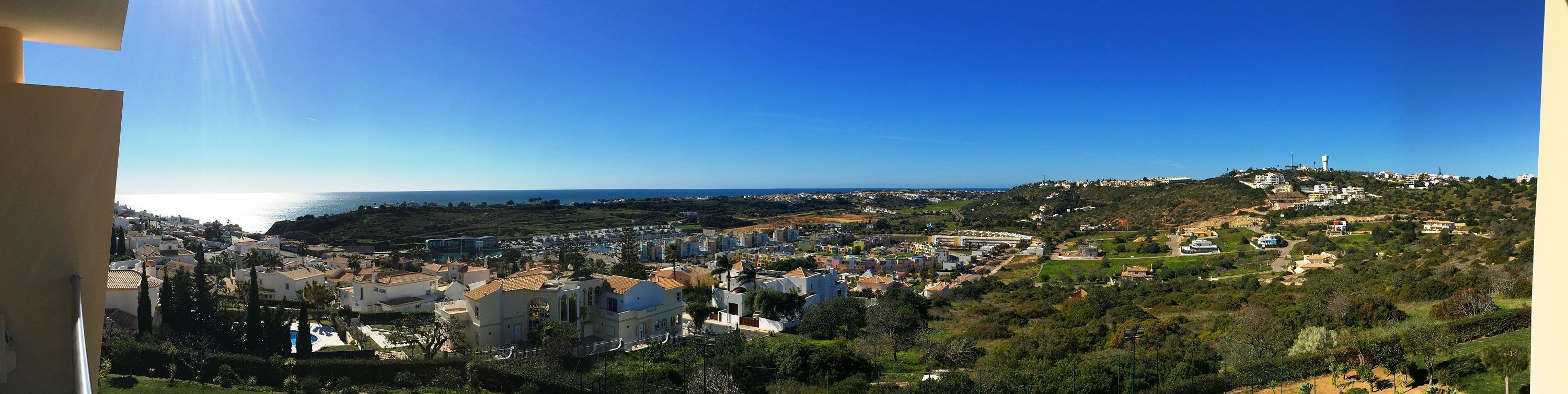 Une vue panoramique depuis un balcon, donnant sur une ville côtière avec ses bâtiments, sa végétation et la mer en arrière-plan.