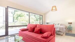 A modern room with a red sofa, a glass coffee table, a bed with a white duvet, and a large window with a view of greenery. - Departamento en Bariloche con Terraza (San Carlos de Bariloche)