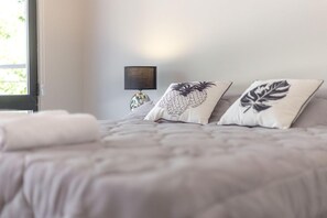 Ein ordentlich bezogenes Bett mit einer grauen Daunendecke, zwei Zierkissen mit Ananas- und Blätter-Motiven und ein kleines gerahmtes Bild an der Wand.
