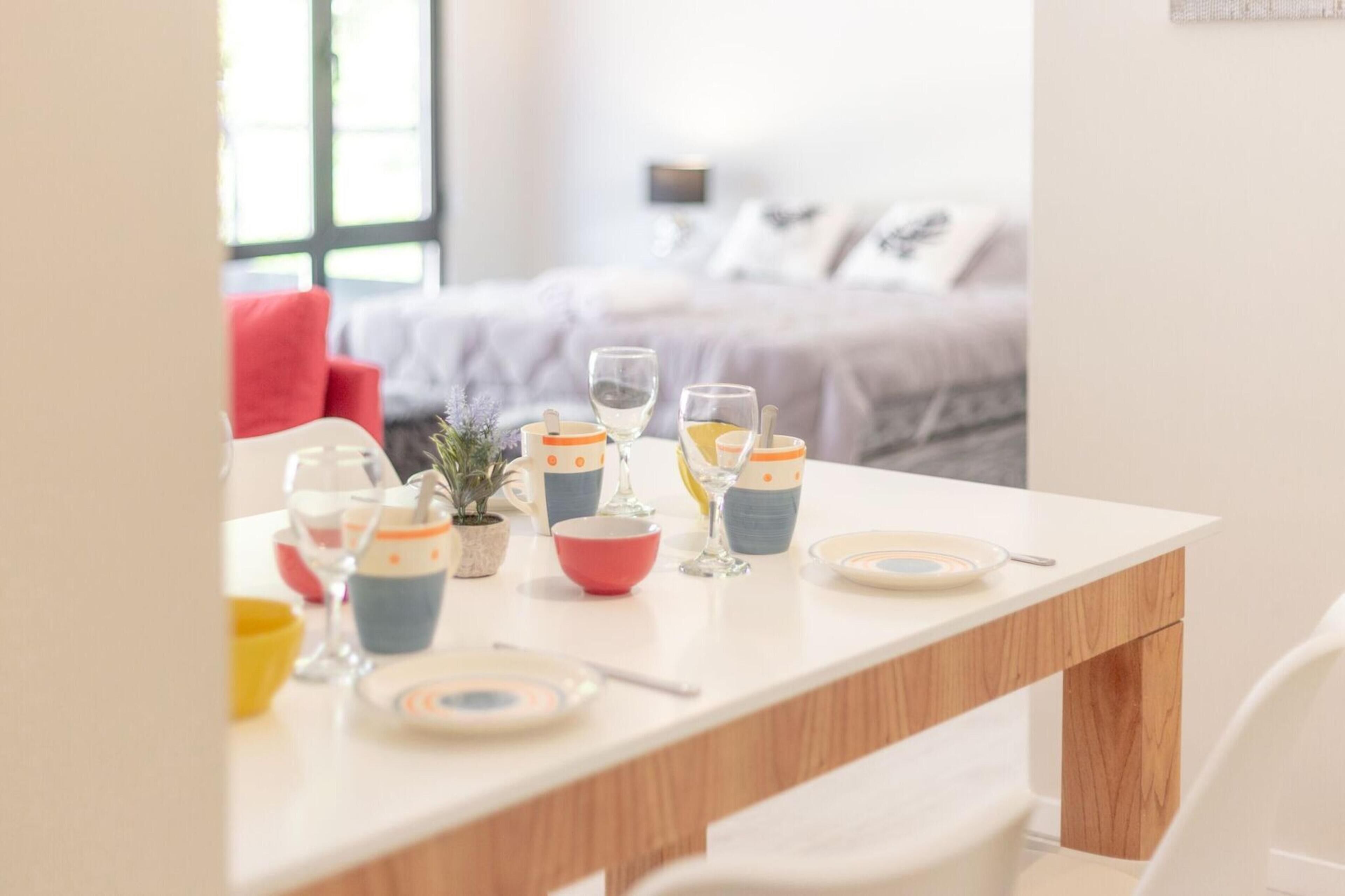 A dining table set with glasses, bowls, and plates, with a bed and pillows in the background.