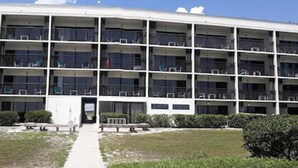 A multi-story building with a central entrance, surrounded by a grassy area and small bushes. - Surf Suites (Wrightsville Beach)