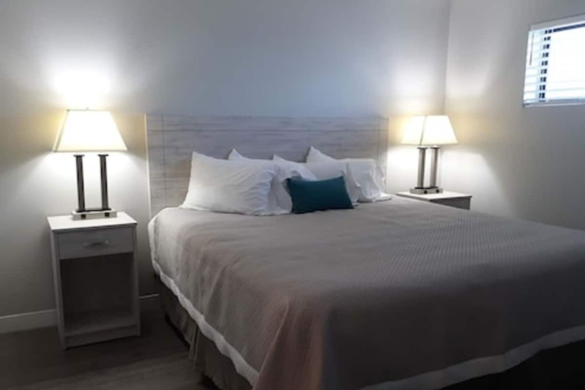 A neatly made bed with white and teal pillows, flanked by two bedside tables with lamps, in a modern room with a neutral color scheme.