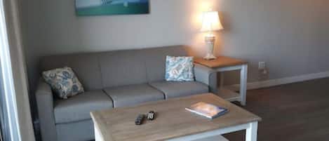 A living room with a grey sofa, a wooden coffee table, a lamp, and a painting of a pier on the wall.