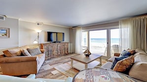 A living room with a sofa, a coffee table, and a television. There is a view of the beach through the glass doors.