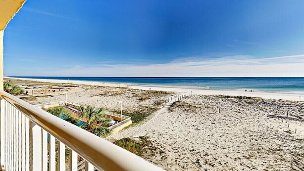 A beach view from a balcony with a clear sky, sandy beach, and some vegetation.