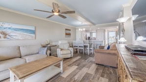 A spacious living room with a large sofa, a coffee table, and a dining area with chairs and a table. A ceiling fan and a painting of a beach scene are visible.