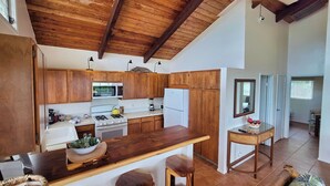 Una cocina con armarios de madera, estufa, microondas y refrigerador. Hay una isla de madera con taburetes y una mesita con un tazón de fruta.