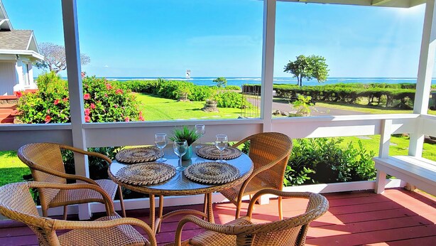 Un espacio exterior techado con sillas de mimbre y una mesa, con vistas a un jardín cuidado y al océano.